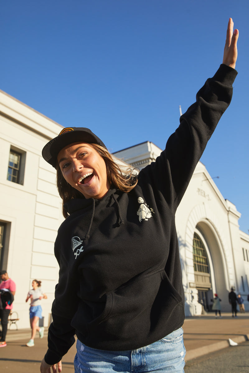 A smiling woman in a black Moon Bridge Pullover Hoodie raises her arm outside a white building on a sunny day.