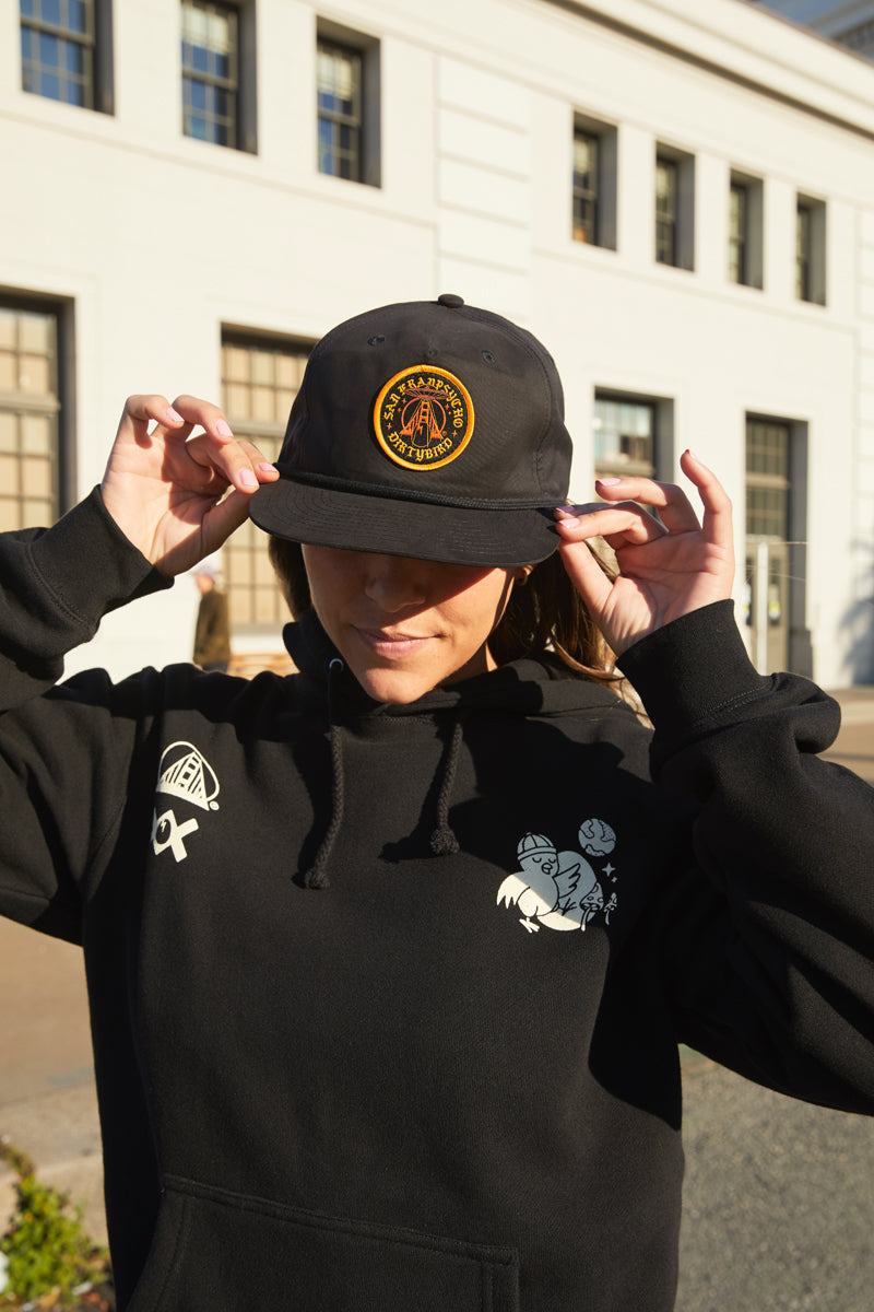 A woman in a black hoodie and Dirty UFO Hat stands outside, adjusting her cap with a white building behind her—a nod to Bay Area culture.