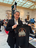 A man in a suit smiles while holding a Let's Go San Francisco Tee at an indoor market or fair.