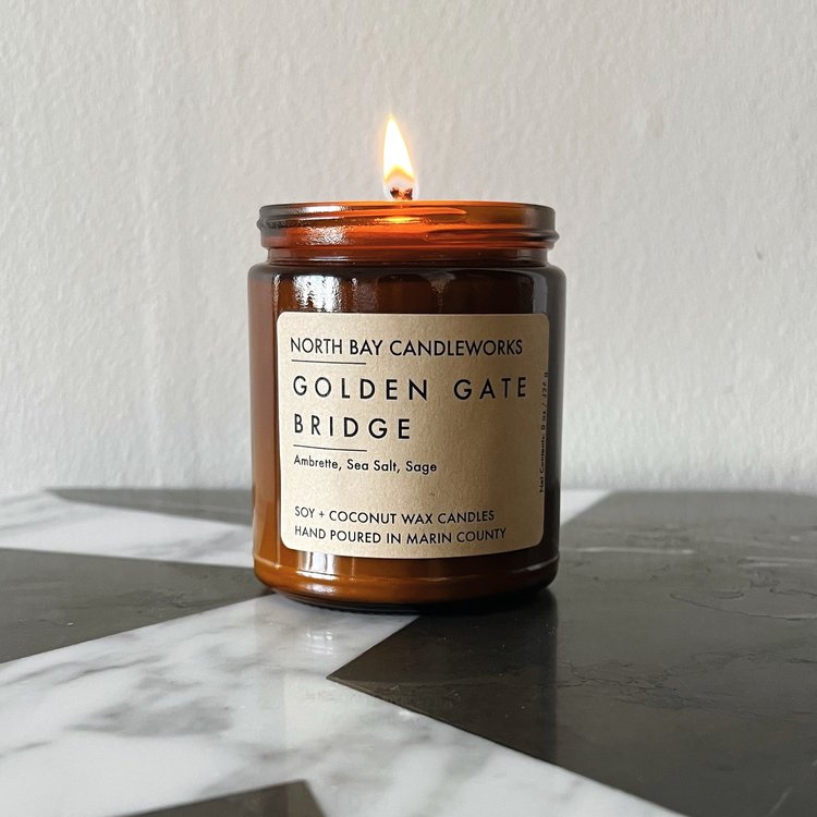 A lit North Bay Candleworks candle in a brown jar sits elegantly on a marble surface, capturing the essence of the Bay Area.