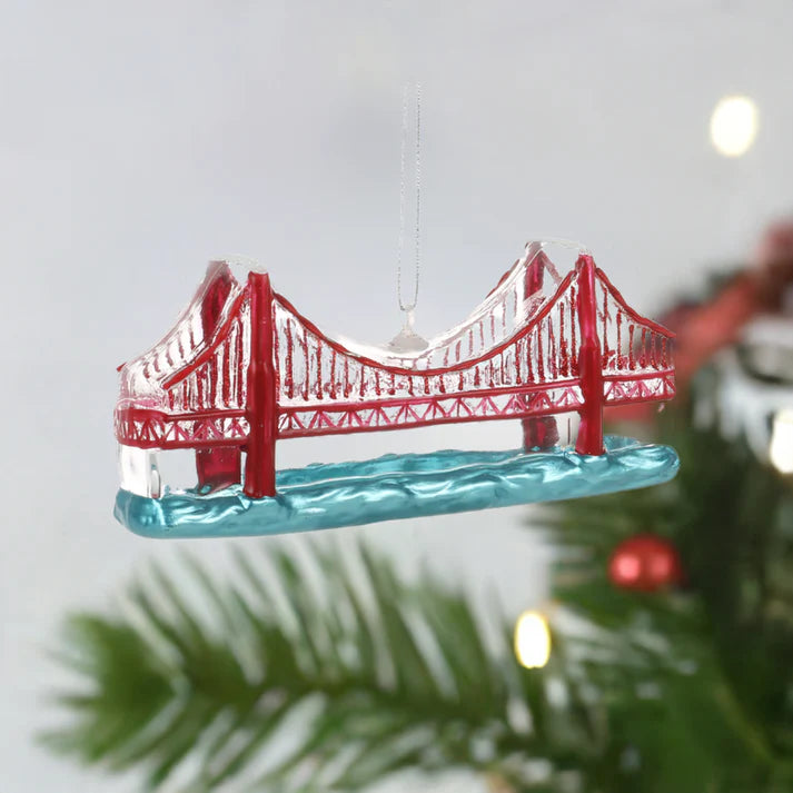 A red suspension bridge-shaped Christmas tree ornament from Local Elves hangs from a branch. This hand-painted glass piece beautifully reflects the city's unique charm.