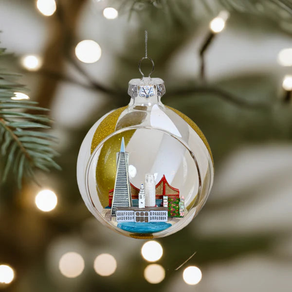 The Xmas Ornaments - Local Elves features a hand-painted glass cityscape, hanging elegantly on a Christmas tree with blurred lights, showcasing the craftsmanship of San Francisco in its durable glass construction.