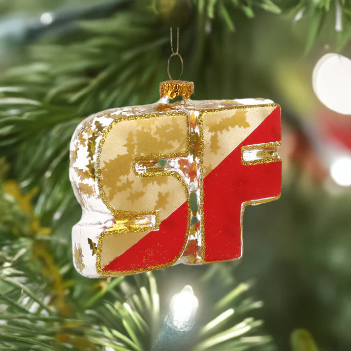 A Xmas Ornament from Local Elves, featuring hand-painted glass with SF in gold and red, hangs gracefully on a Christmas tree.