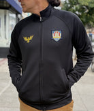 Person wearing the WC x SFP Zip-Up Jacket, a black jacket with gold and colorful embroidered logos, standing outdoors with hands in pockets.