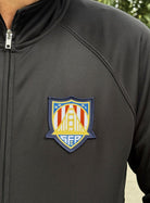 Person wearing the WC x SFP Zip-Up Jacket in black, featuring a colorful shield patch with a bridge and geometric designs.