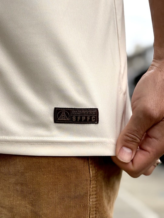 A hand holds the hem of a cream WC x SFP Jersey - Away with an SFPFC patch, next to brown corduroy pants.
