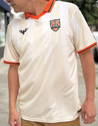 A man is wearing the WC x SFP Jersey - Away, a cream fan-fit top with orange trim and two chest logos, inspired by classic World Cup jerseys, while standing outdoors.