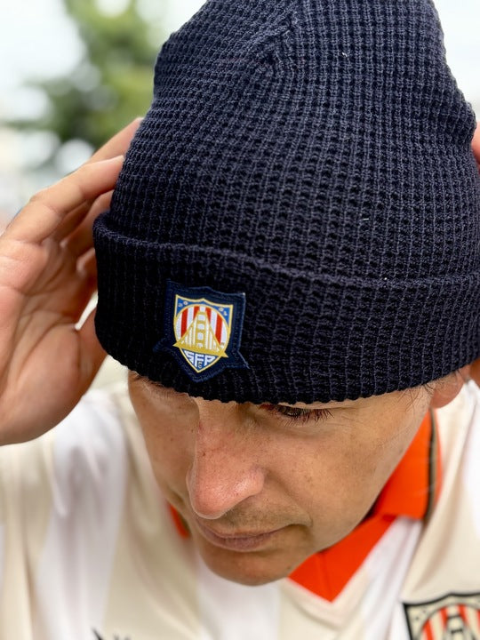 Man in a white shirt and black beanie featuring a crest, looking down and adjusting his WC x SFP Beanie with both hands.