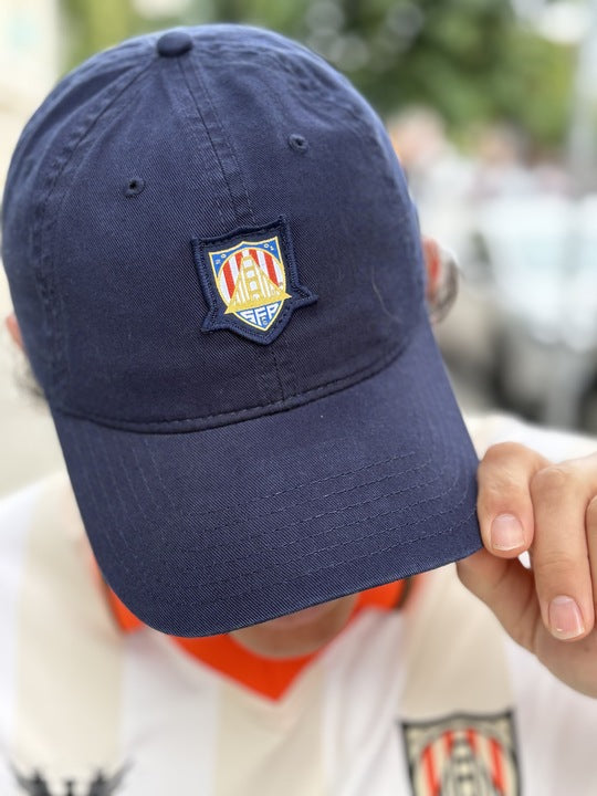 Individual wearing a WC x SFP Patched Hat in navy blue featuring the US Men's National Team patch, paired with a white-striped shirt showcasing the matching shield emblem, while holding the hat’s brim.