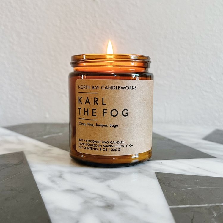 A lit amber candle from North Bay Candleworks, labeled Karl the Fog, sits on a marble surface, capturing the mysterious allure of the Bay Area.