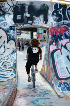 A person with curly hair rides a bike through a graffiti-covered concrete archway in an urban skate park, wearing the Black & Orange Street Skull Tee made from soft Airlume cotton.
