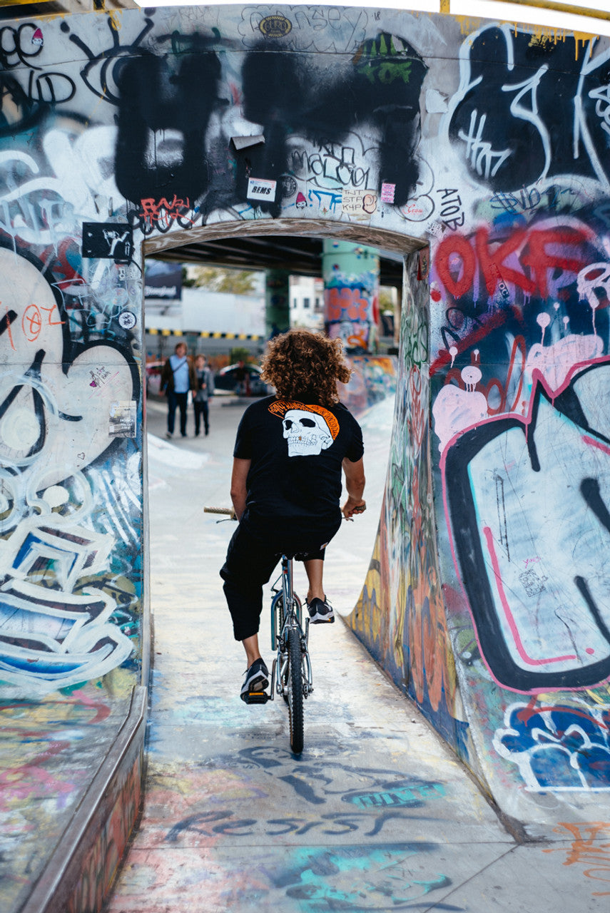 A person with curly hair rides a bike through a graffiti-covered concrete archway in an urban skate park, wearing the Black & Orange Street Skull Tee made from soft Airlume cotton.