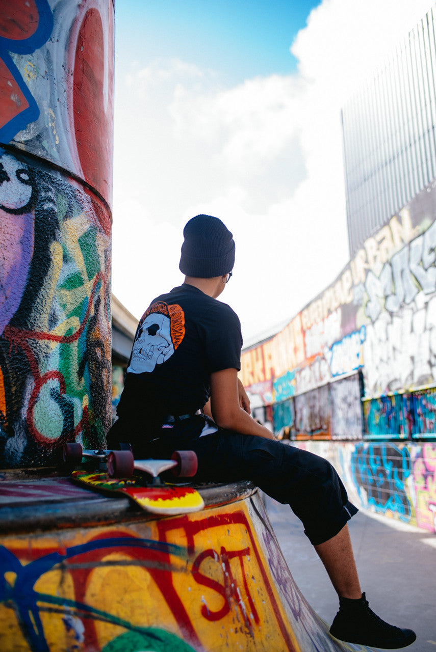 A person sits on a skateboard in a graffiti-covered skate park, wearing a beanie and a Black & Orange Street Skull Tee made from soft Airlume cotton.