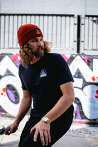 A man with curly hair, donning a red beanie and a Black & Orange Street Skull Tee, stands by a graffiti-covered wall.