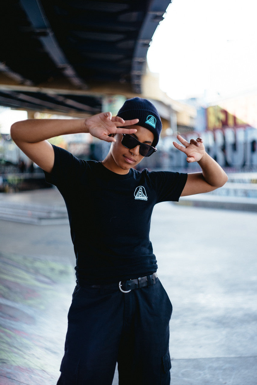 A person wearing the Black & Orange Street Skull Tee and sunglasses poses under an overpass, flashing peace signs with both hands. The hand-printed design on the Airlume cotton enhances their cool, effortless style with a unique touch.