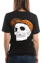 The person is wearing the Black & Orange Street Skull Tee, distinguished by its bold black design, prominently displaying a large hand-printed skull on the back with vibrant orange text encircling it. Made from soft Airlume cotton, this shirt seamlessly blends comfort with an edgy style.