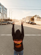 A person in an Outer Sunset Zip-Up Hoodie stands triumphantly on the empty expanse of Noriega Street, their arms raised high. Buildings cast long shadows over parked cars beneath a bright, sunny sky.