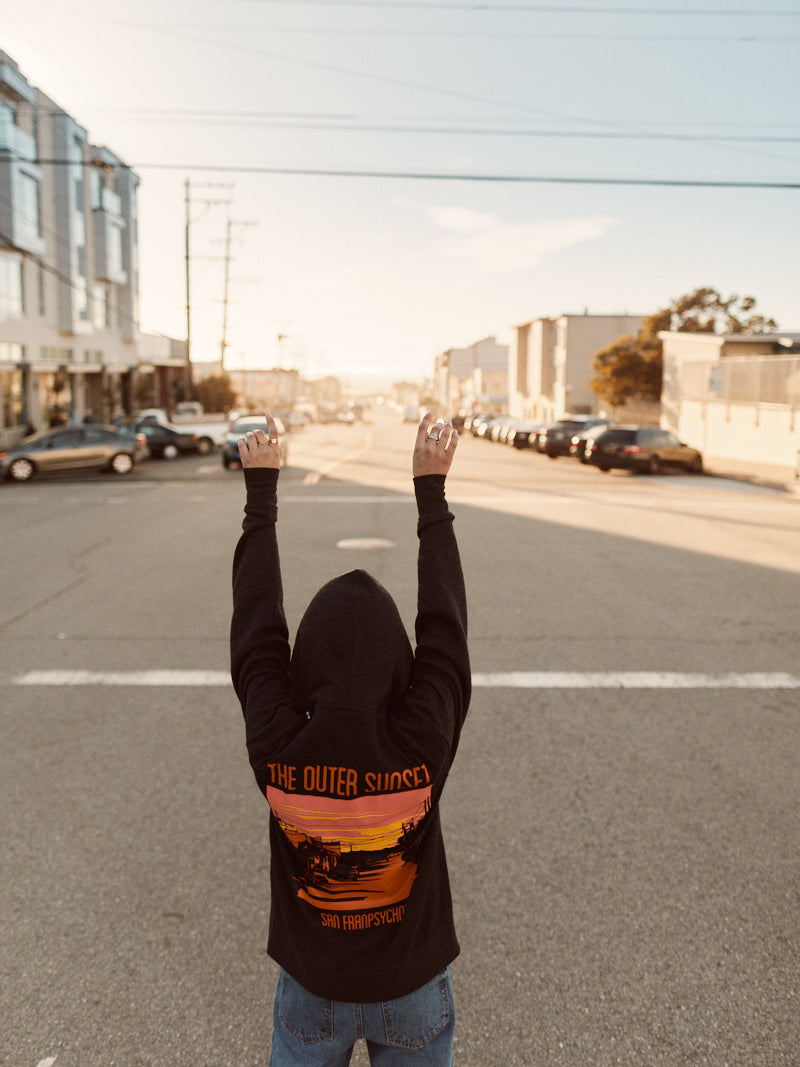 A person in an Outer Sunset Zip-Up Hoodie stands triumphantly on the empty expanse of Noriega Street, their arms raised high. Buildings cast long shadows over parked cars beneath a bright, sunny sky.