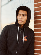 A person wearing an Outer Sunset Zip-Up Hoodie leans against a brick wall on Noriega St, gazing thoughtfully into the distance.