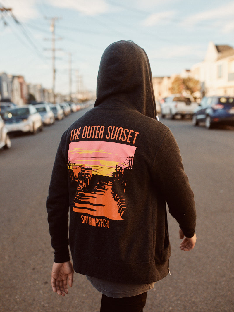 A person strolls down Noriega St, sporting the Outer Sunset Zip-Up Hoodie with a vibrant sunset graphic and "The Outer Sunset, San Francisco" text.