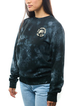 A person wearing the Surf Club Crewneck, a black tie-dye, super-soft sweatshirt featuring "Skeleton Surf Club" text, paired with light blue jeans, standing against a white background.