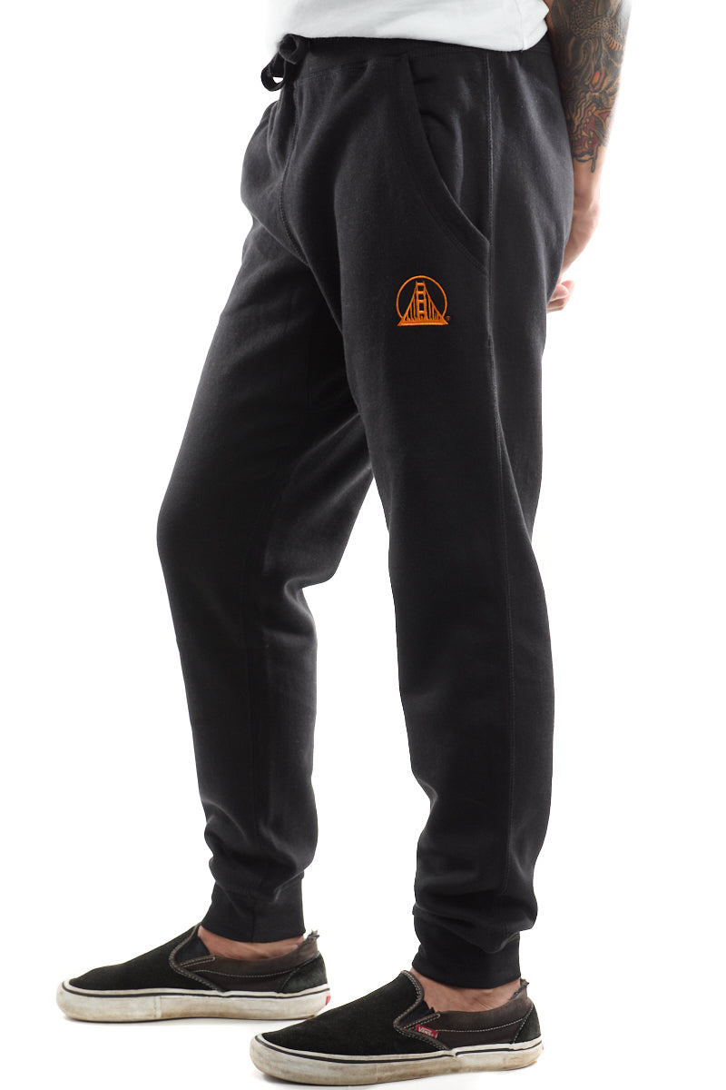 Dressed in Black & Orange Embroidered Logo Joggers, the individual stands comfortably in sleek black slip-on shoes.