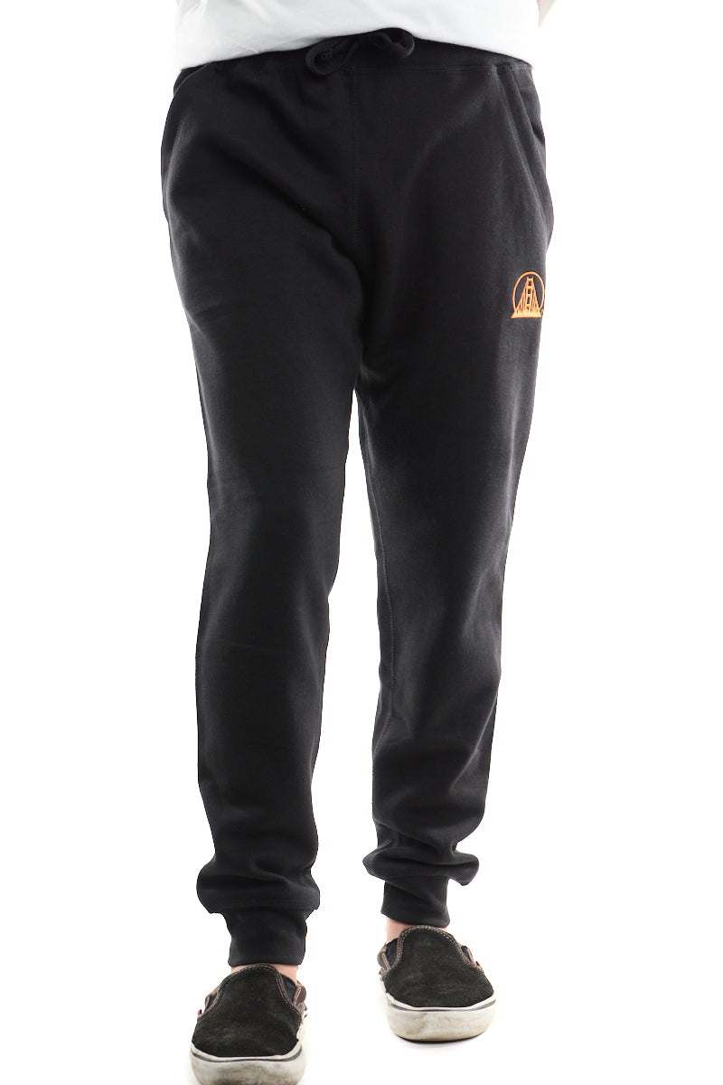 A person donning the Black & Orange Embroidered Logo Joggers, with a small orange emblem near the pocket, paired with comfortable black slip-on shoes, embodies casual elegance.