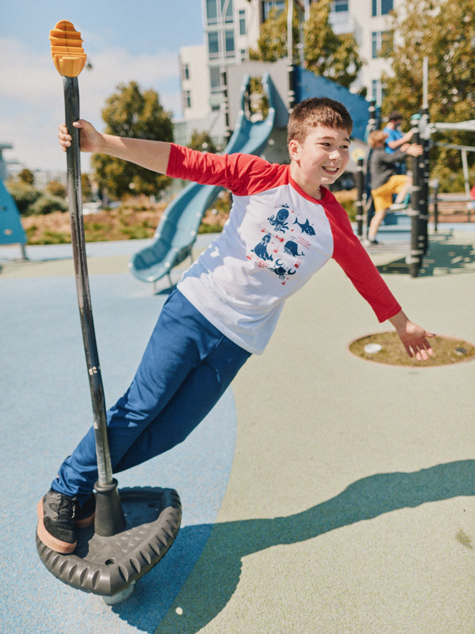 A boy in a Youth Ocean Club Baseball Tee, made from 100% cotton, enjoys a spinning ride at a sunny playground.