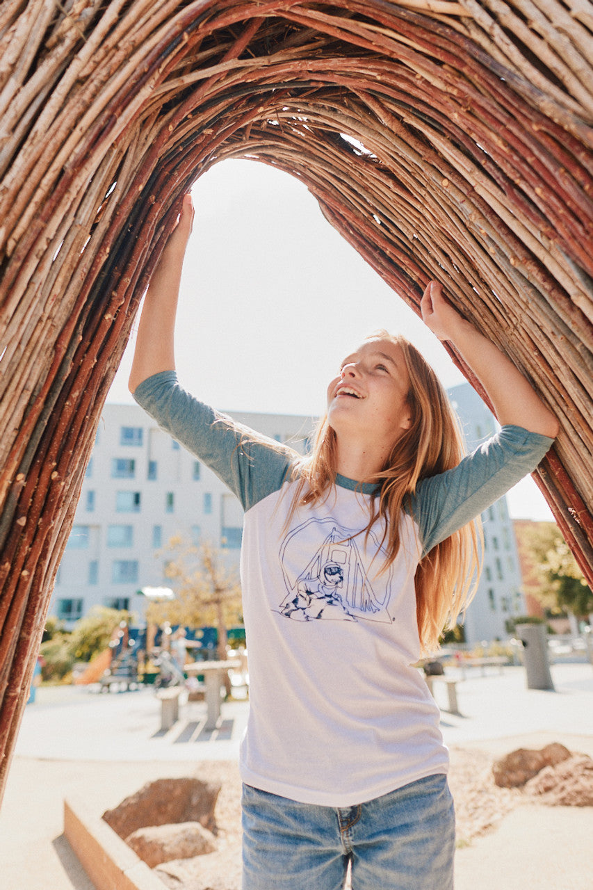 In a sunny outdoor setting, a person joyfully stands under a wooden arch structure, sporting the vibrant Youth Seal Logo Baseball Tee crafted from 100% cotton.