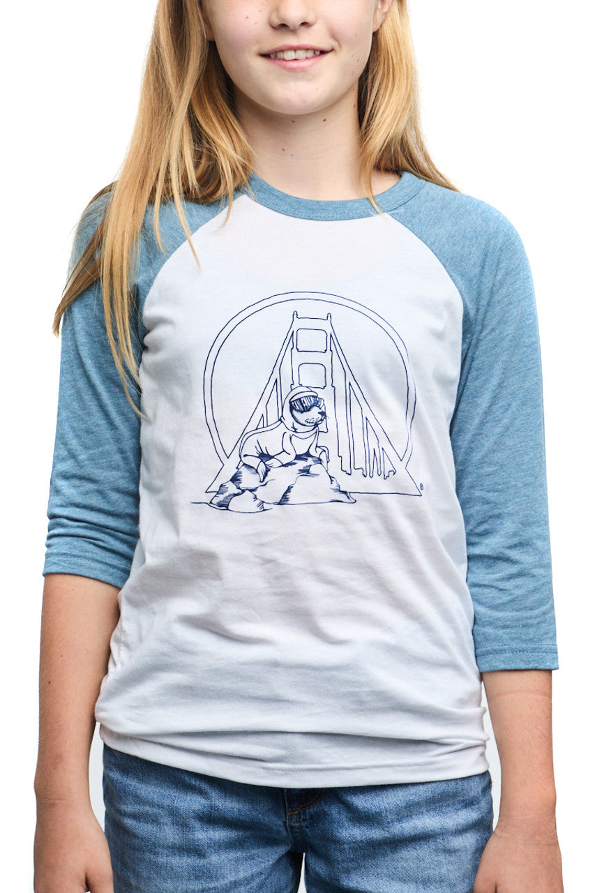 A cheerful girl is captured smiling at the camera, wearing a Youth Seal Logo Baseball Tee in blue and white, crafted from 100% cotton and featuring a charming bridge design that's perfect for kids.