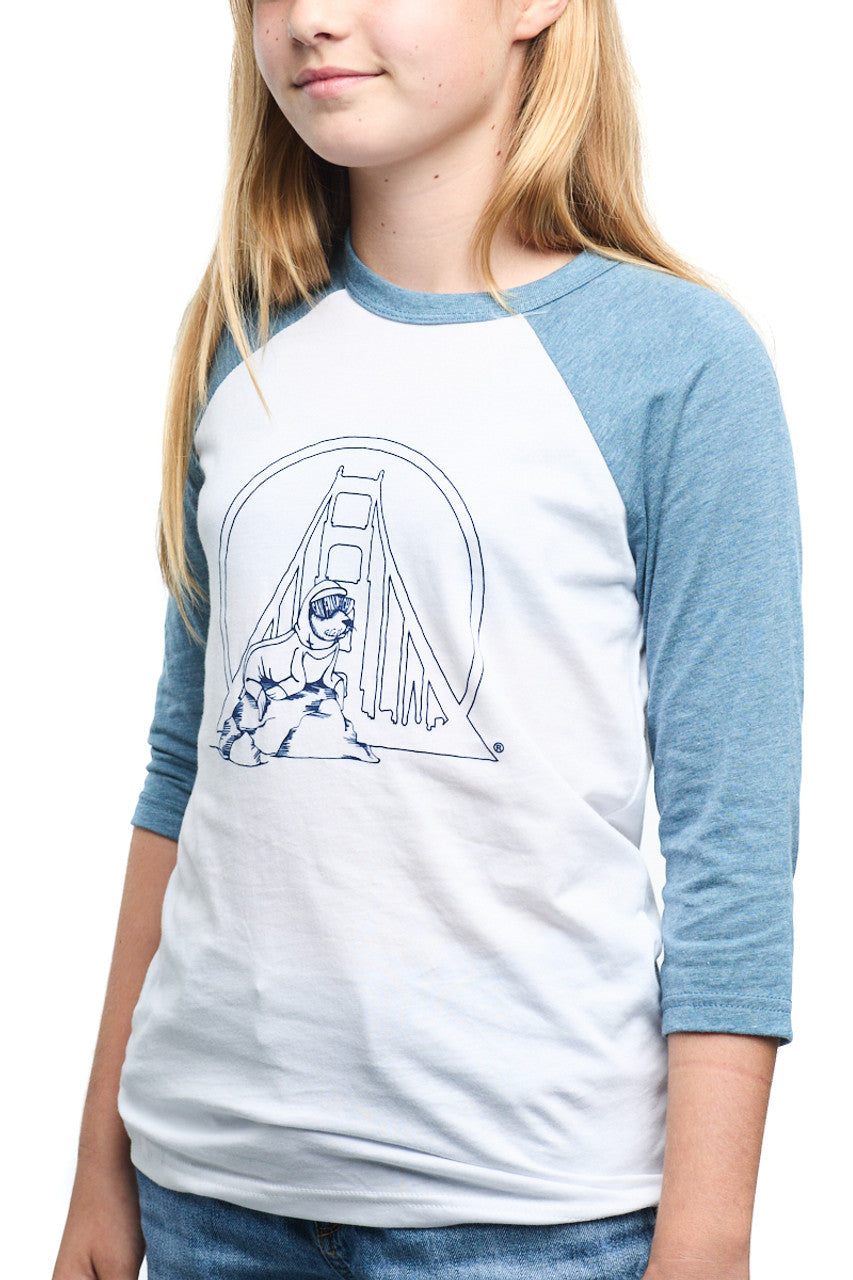 A girl wearing the trendy Youth Seal Logo Baseball Tee, made from 100% cotton, showcasing a blue and white design with a graphic of a bridge and person on the front. Ideal for kids who appreciate comfort and style.