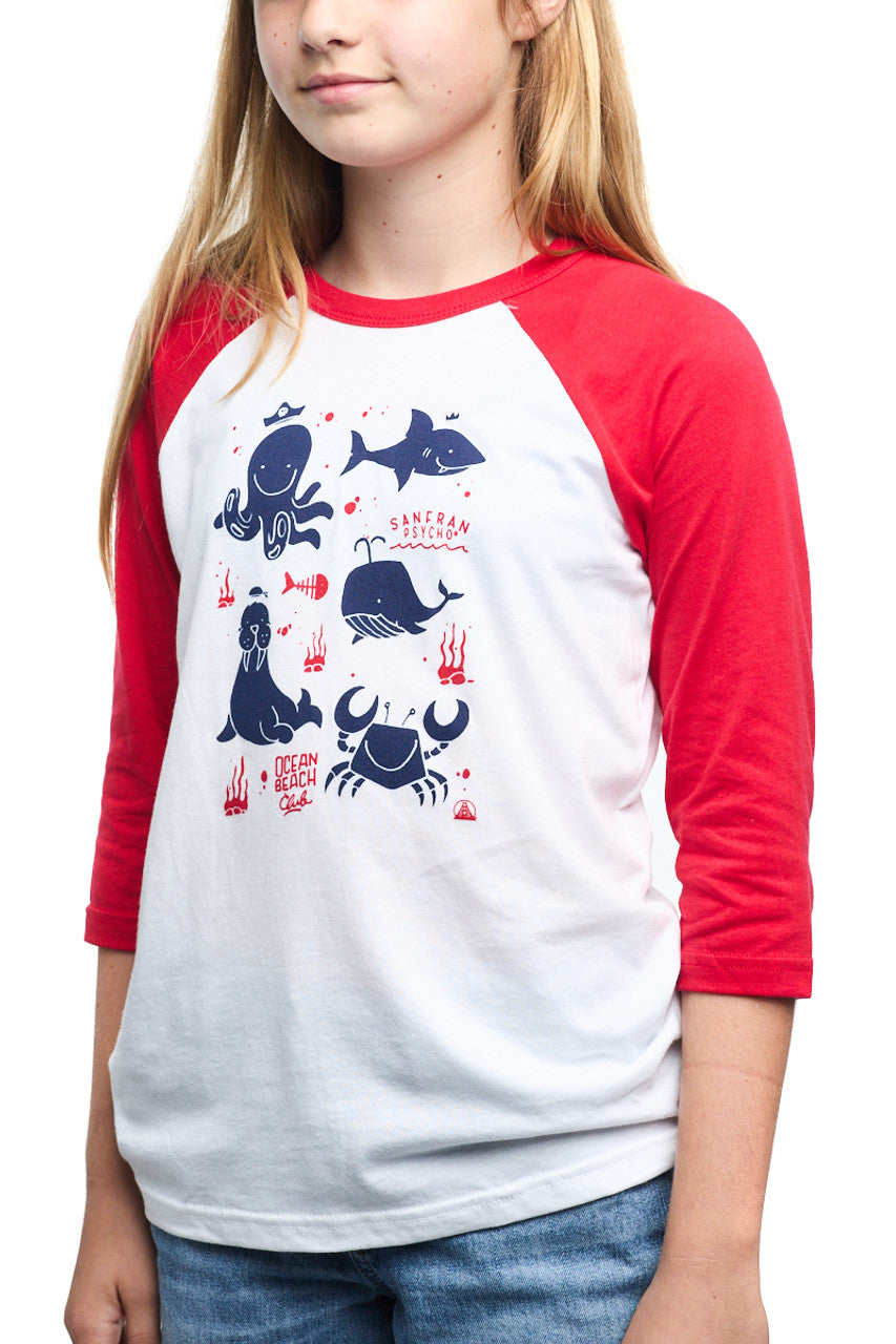 A child wears the Youth Ocean Club Baseball Tee featuring vibrant red sleeves, playful ocean animal illustrations, and the Kids Ocean Club label. Made from 100% cotton, it merges comfort with style.