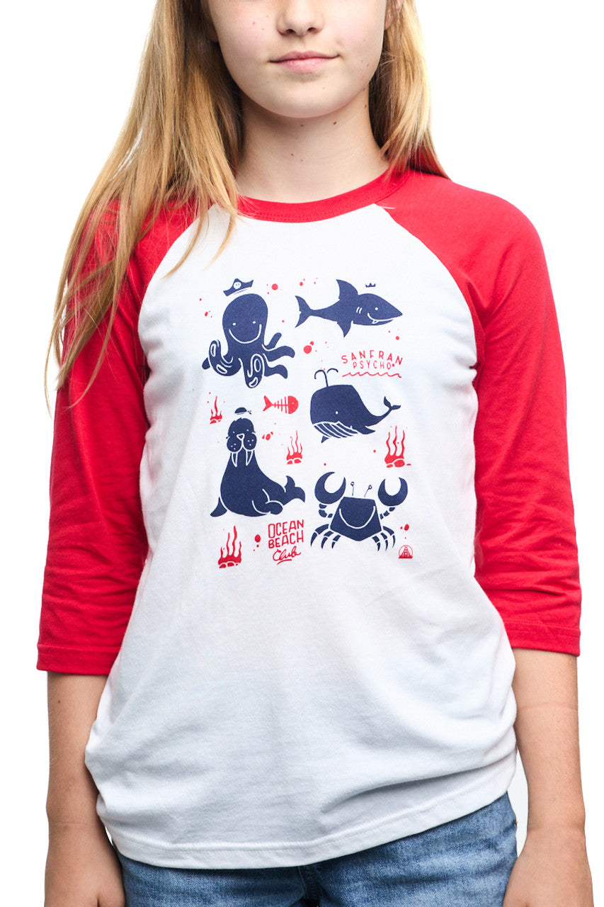 A girl wears the Youth Ocean Club Baseball Tee, featuring red and white colors and marine animal illustrations. Made from 100% cotton, it offers comfort and style for young Kids Ocean Club members.