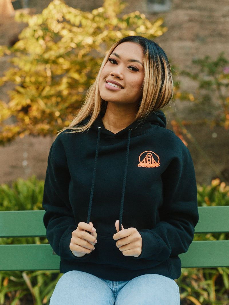 A person sits on a bench in San Francisco, wearing the Black & Orange Logo Pullover, against the vibrant backdrop of autumn foliage.
