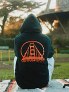 A person sits outdoors in a Black & Orange Logo Pullover by San Franpsycho, showcasing a striking bridge graphic on the back that perfectly captures the spirit of San Francisco.