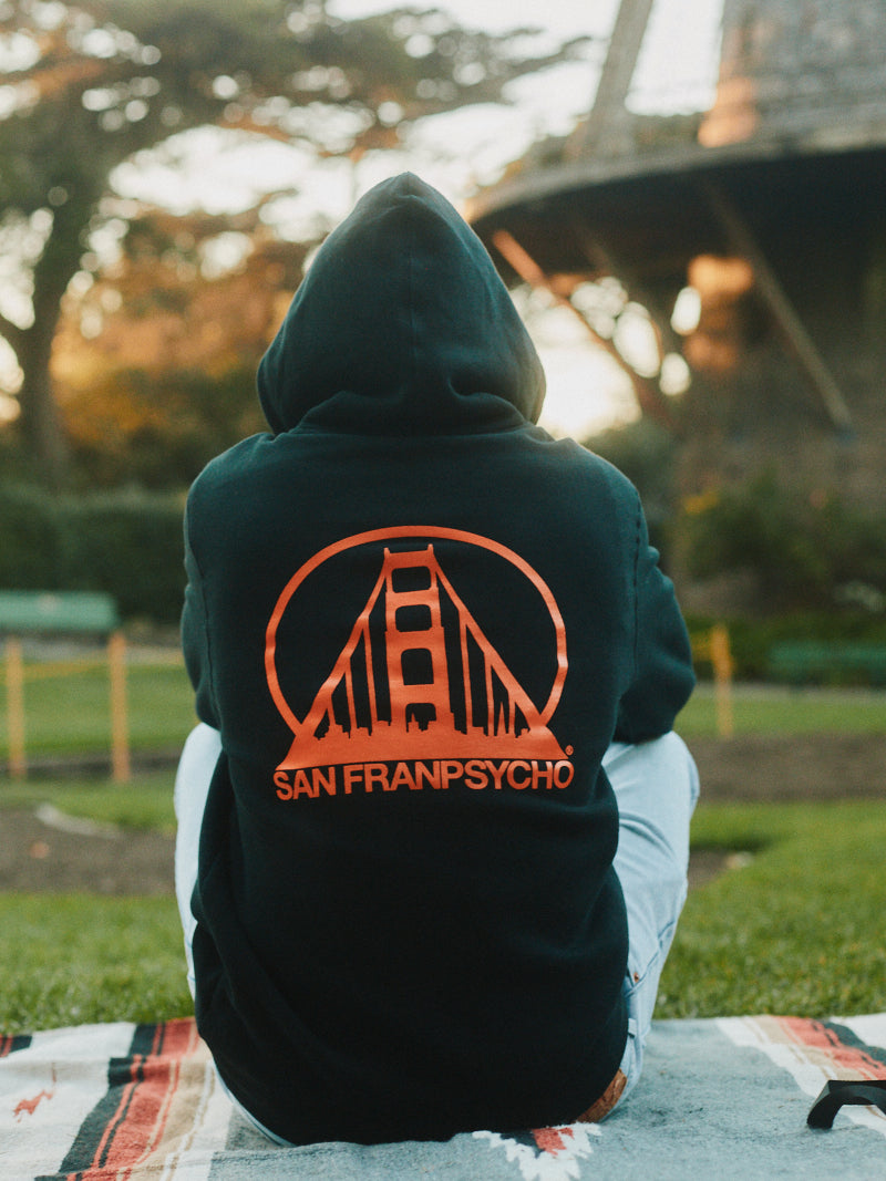 A person sits outdoors in a Black & Orange Logo Pullover by San Franpsycho, showcasing a striking bridge graphic on the back that perfectly captures the spirit of San Francisco.