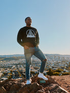 A man wearing the Black & White Logo Crewneck and jeans stands on a rock, with a cityscape sprawling beneath the clear sky. The iconic SFP logo subtly adorns his attire, embodying elegance and versatility, available in various size options.