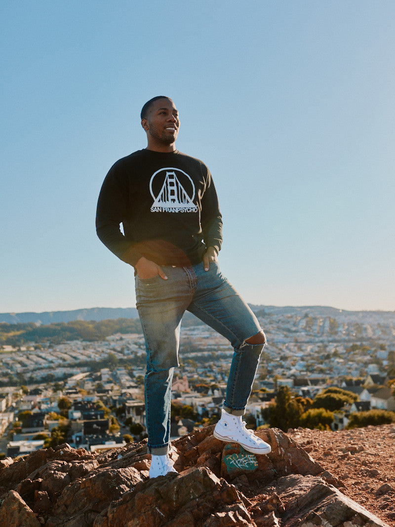A man wearing the Black & White Logo Crewneck and jeans stands on a rock, with a cityscape sprawling beneath the clear sky. The iconic SFP logo subtly adorns his attire, embodying elegance and versatility, available in various size options.