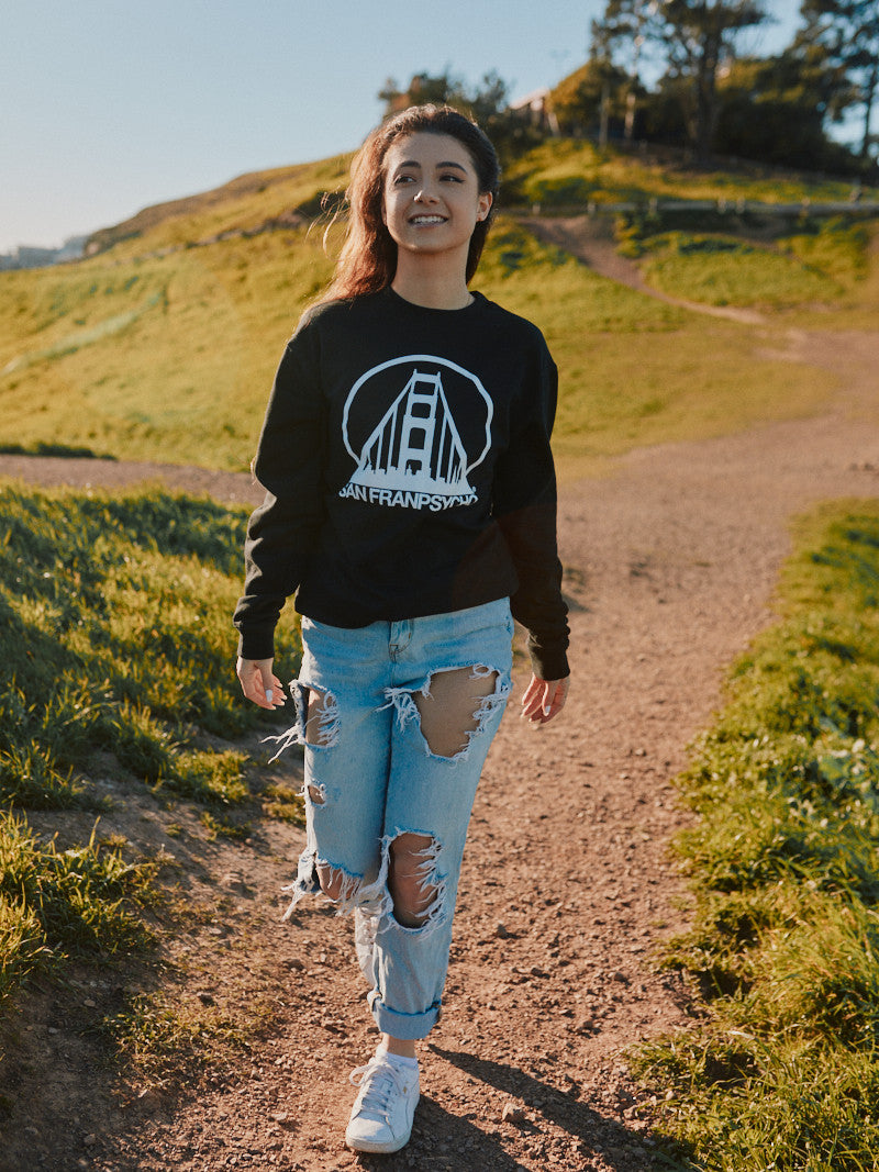 A person strolls down a sunny path wearing the Black & White Logo Crewneck and ripped jeans, with hills and trees painting the backdrop. The sleek black fabric gracefully displays the subtle SFP logo, available in various sizes to ensure the perfect fit.
