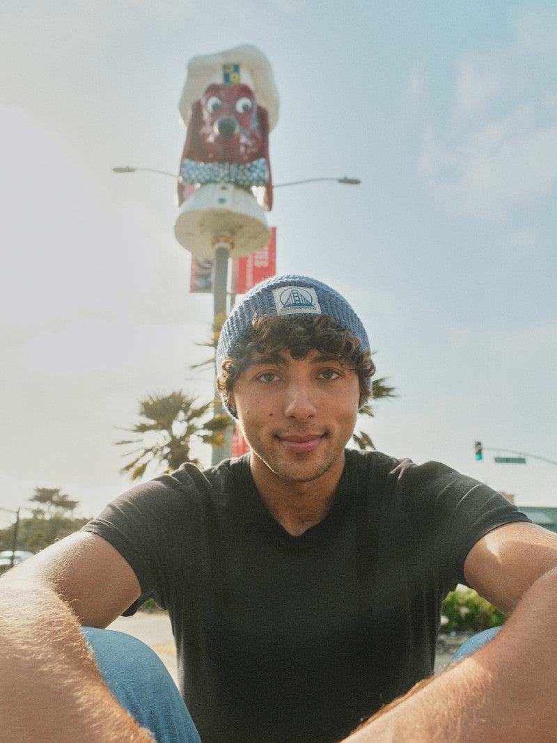 A young man wearing the Denim Heather Waffle Beanie with a Natural/Navy SFP Logo smiles while sitting outdoors under a sunny sky, with a whimsical character statue behind him.