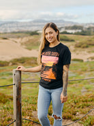 A person with long hair stands outdoors by a fence, wearing an Outer Sunset Tee from San Franpsycho that features a colorful sunset design capturing the essence of San Francisco's Outer Sunset.