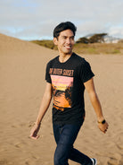A person strolls through a sandy landscape, wearing the Outer Sunset Tee with a sunset print. Smiling and looking away, they embody the laid-back spirit of San Franpsycho, reflecting the Outer Sunset vibes of San Francisco.