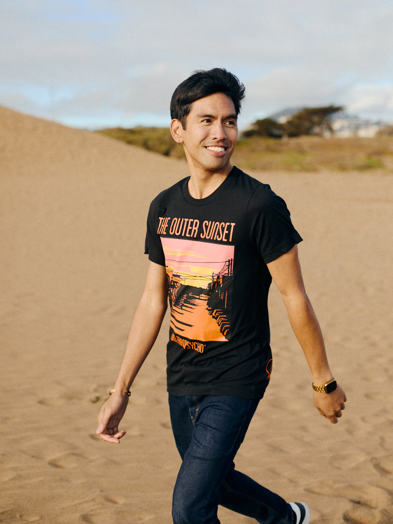 A person strolls through a sandy landscape, wearing the Outer Sunset Tee with a sunset print. Smiling and looking away, they embody the laid-back spirit of San Franpsycho, reflecting the Outer Sunset vibes of San Francisco.