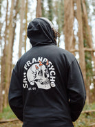 A person wearing a Shroom Skull Zip-Up Hoodie, featuring SAN FRANPSYCHO text, stands mysteriously in the forest.