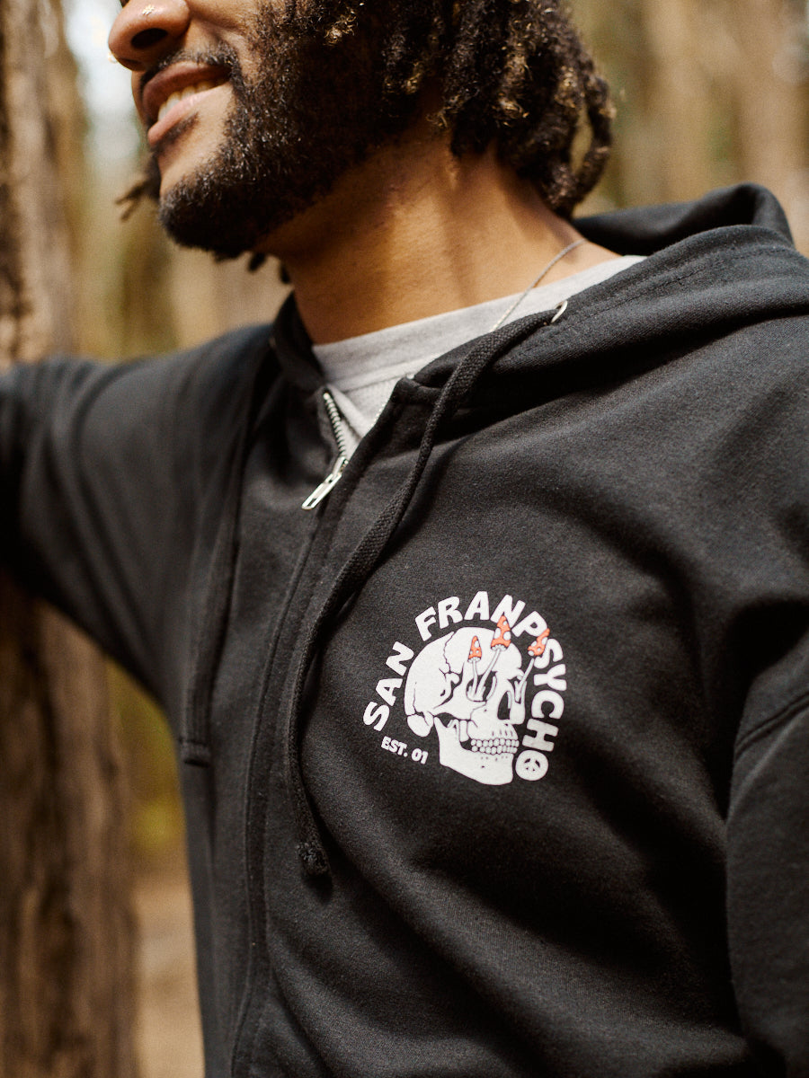 Wearing a black Shroom Skull Zip-Up Hoodie, made from 100% cotton and featuring a unique hand-printed design, a person smiles while standing in a forest.