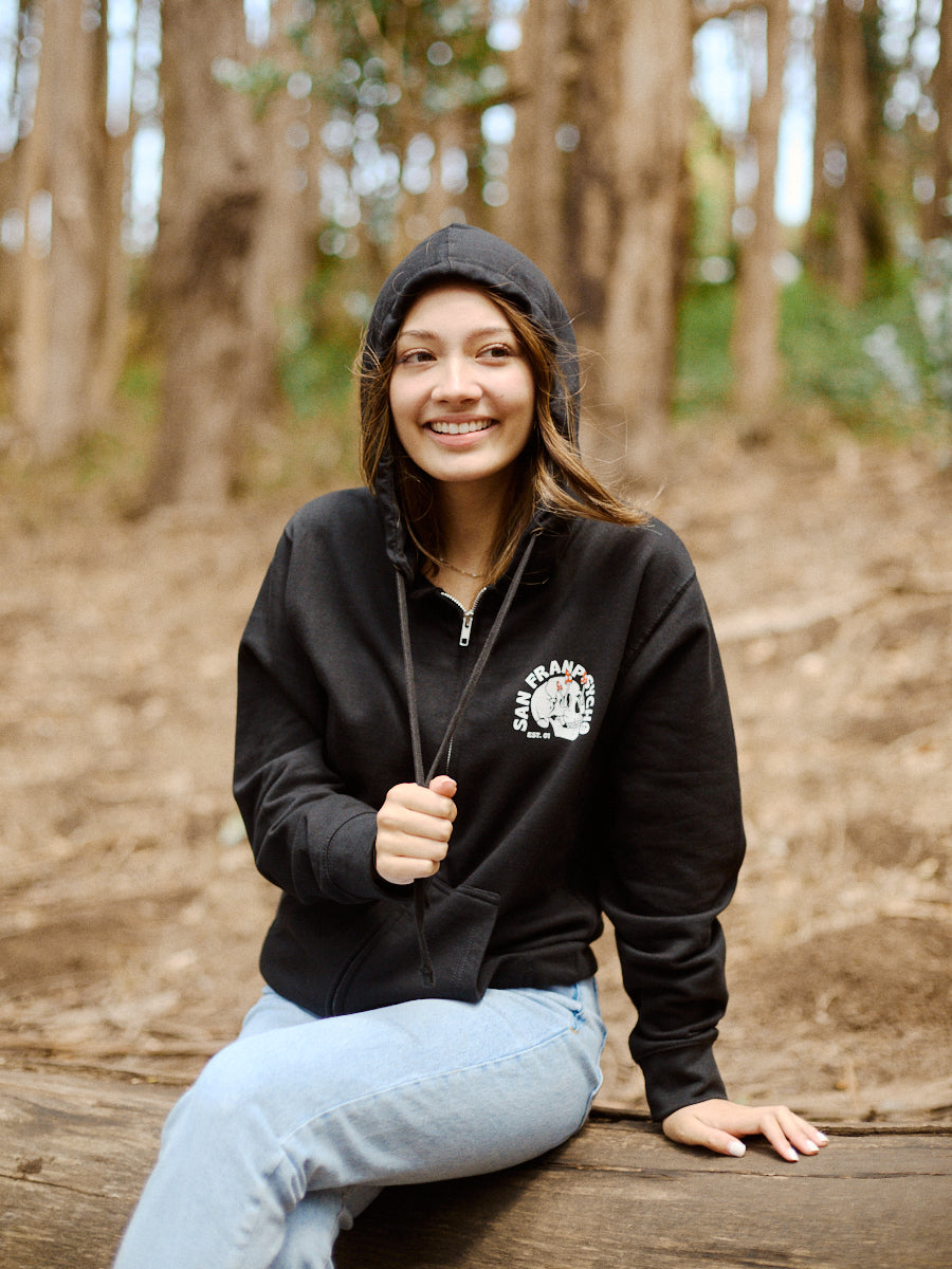 A person wearing a Shroom Skull Zip-Up Hoodie, made from 100% cotton, sits on a log in the forest, smiling while holding onto the zipper.