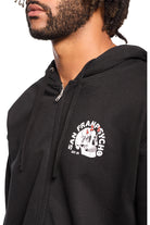 The individual is wearing a black Shroom Skull Zip-Up Hoodie from San Franpsycho, adorned with a graphic logo on the chest. Made entirely of 100% cotton, this piece provides premium comfort and style, highlighting the brand's distinctive hand-printed design.