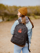 A person in a beanie and jacket, carrying a Black Backpack with a Build Bridges Patch that features leather trims and a red logo, stands outdoors.
