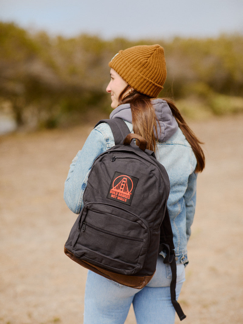 A person in a beanie and jacket, carrying a Black Backpack with a Build Bridges Patch that features leather trims and a red logo, stands outdoors.