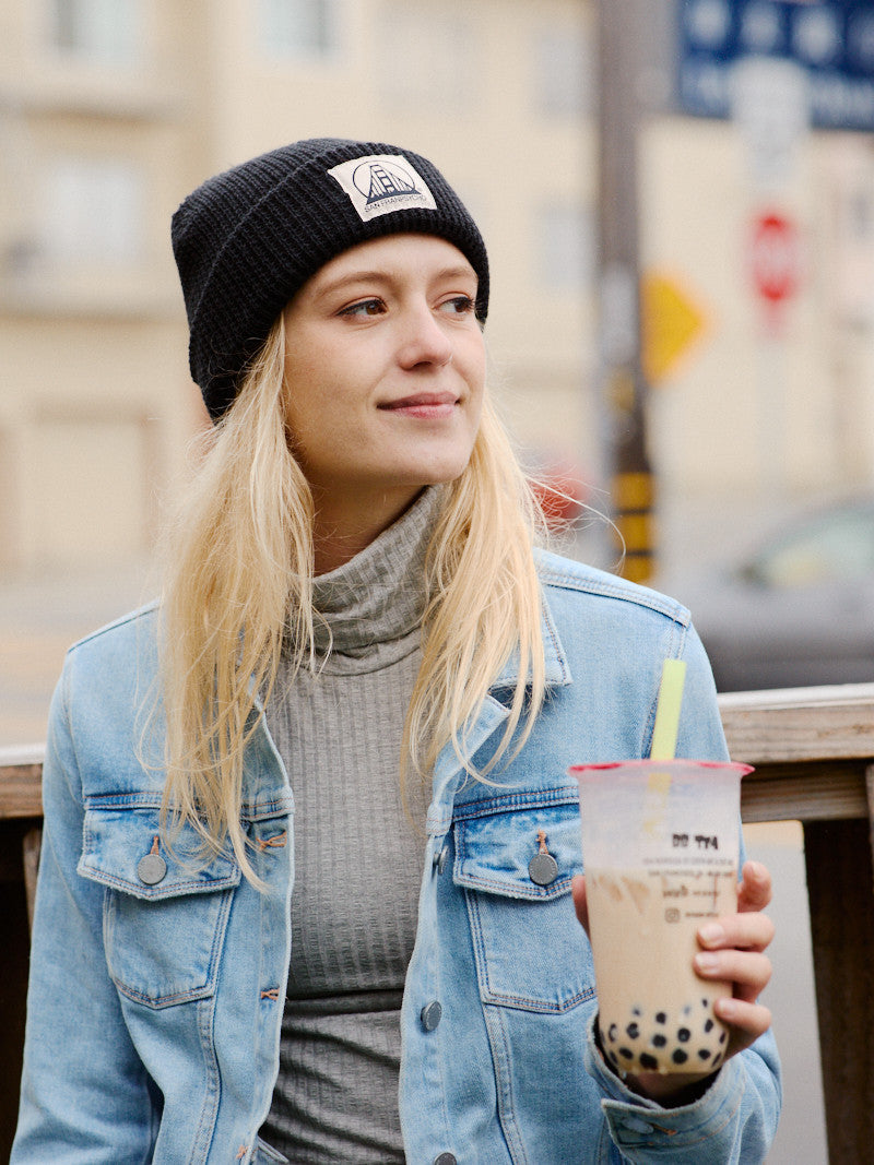 A woman in a denim jacket beams with happiness while wearing the Black Waffle Beanie with Natural/Black SFP Logo and holding a bubble tea outdoors.